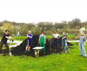 A group of people with a wheelbarrow and shovels stand by a heap of soil.