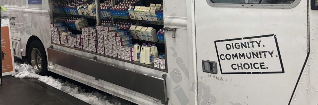 The side of a truck open to reveal carton and bottles of milk. The text Diginity, Community, Choice printed on the side.
