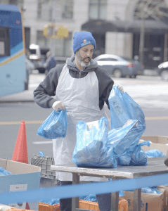 Greg Silverman stands behind a table on the pavement, organising blue plastic bags full of food. 