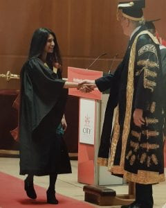 Zara is on stage in her graduation robe and shakes hand with a man in a golden robe. 
