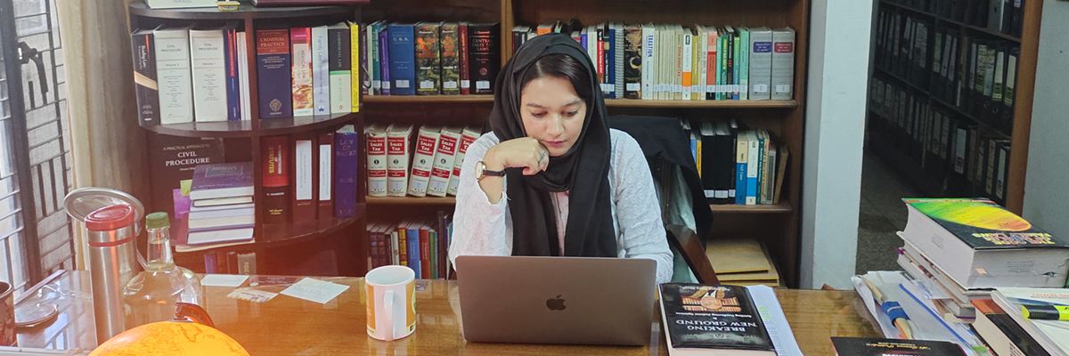 Khadija sits work on a laptop surrounded by books in a large office.