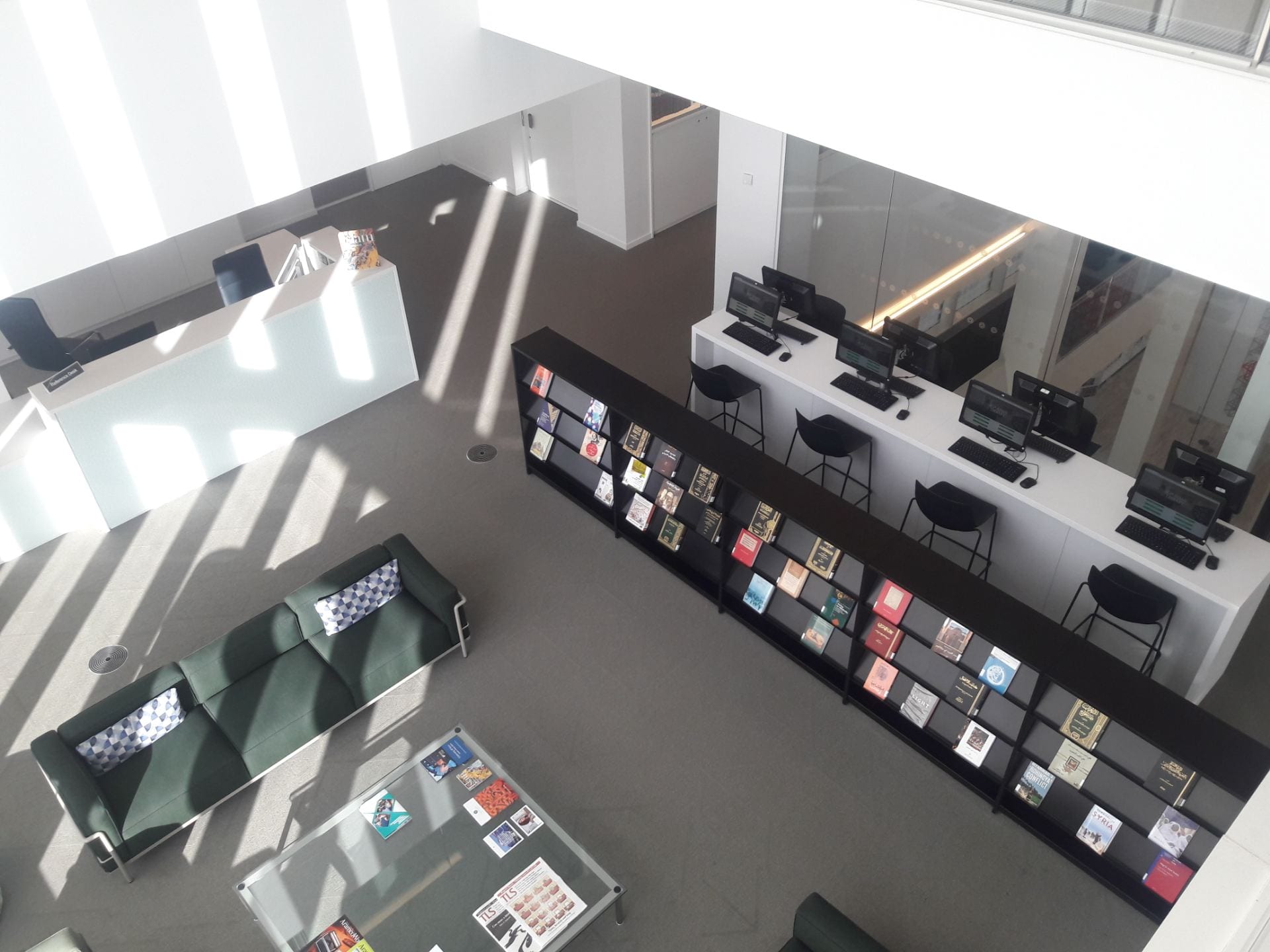 The entrance area of the Aga Khan Library taken from the floor above. The Library Help desk, soft furnishing, coffee table, journal shelves and catalogues and stools can be seen. There is light streaming in.