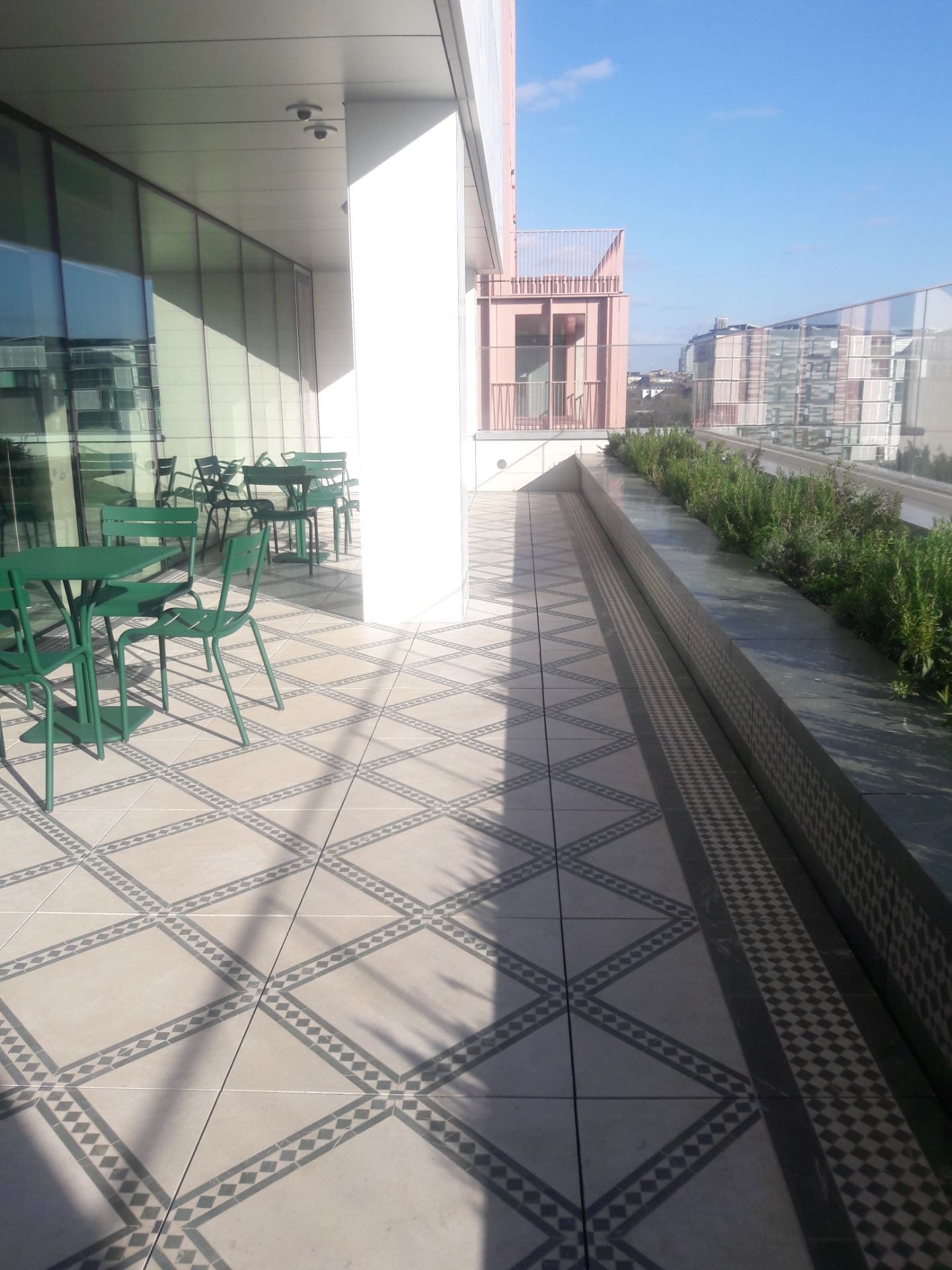 The Terrace of learning, a semi covered terrace in the sunlight. Floor tiles with geometric pattern in the North African style. Green garden tables and chairs sit close to the full length windows. Green evergreen herbs can be seen in the planter.