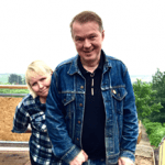 Photo of Edwyn and Grace in front of a field in the countryside. Edwyn stands in front wearing a denim jacket and black top. Grace stands behind him peering around his right shoulder and looking into the camera.