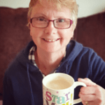 Jan sits on a sofa, smiling up at the camera. She is holding up a mug of tea in her left hand. The mug says the words "Star Mum"