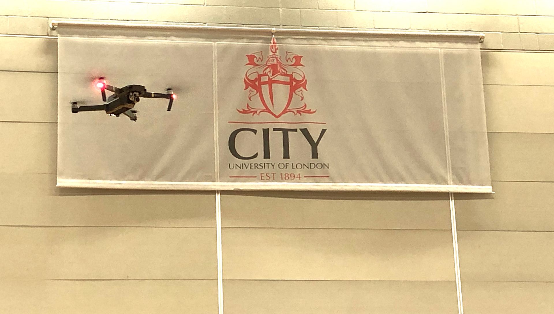 Drone flying in front of City, University of London banner on wall of CitySport gym