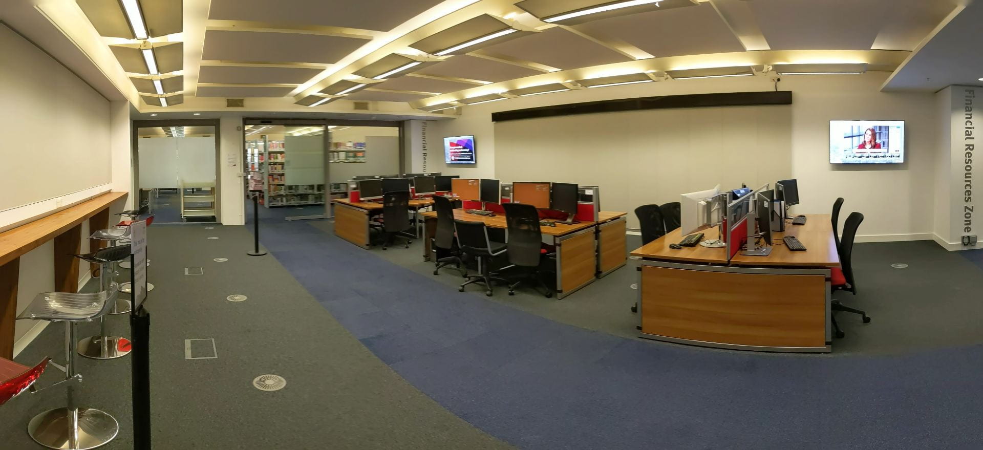 Panoramic image of a library space with three blocks of four computers, underneath a ticker and two video screens. The colour scheme is grey, brown and teal.