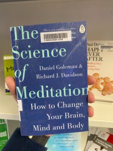 A hand is holding a print copy of 'The Science of Meditation - how to change your brain, mind and body,' by Daniel Goleman and Richard J. Davidson.