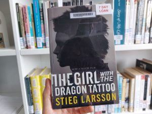A photo of a hand holding a print copy of The Girl with the Dragon Tattoo. Library shelves full of books can be seen in the background